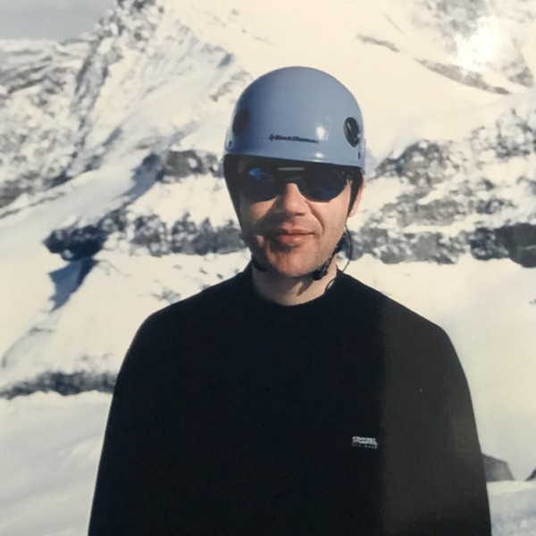 A young Chuck Garcia in a climbing helmet and sunglasses, with snow-covered peaks behind him.