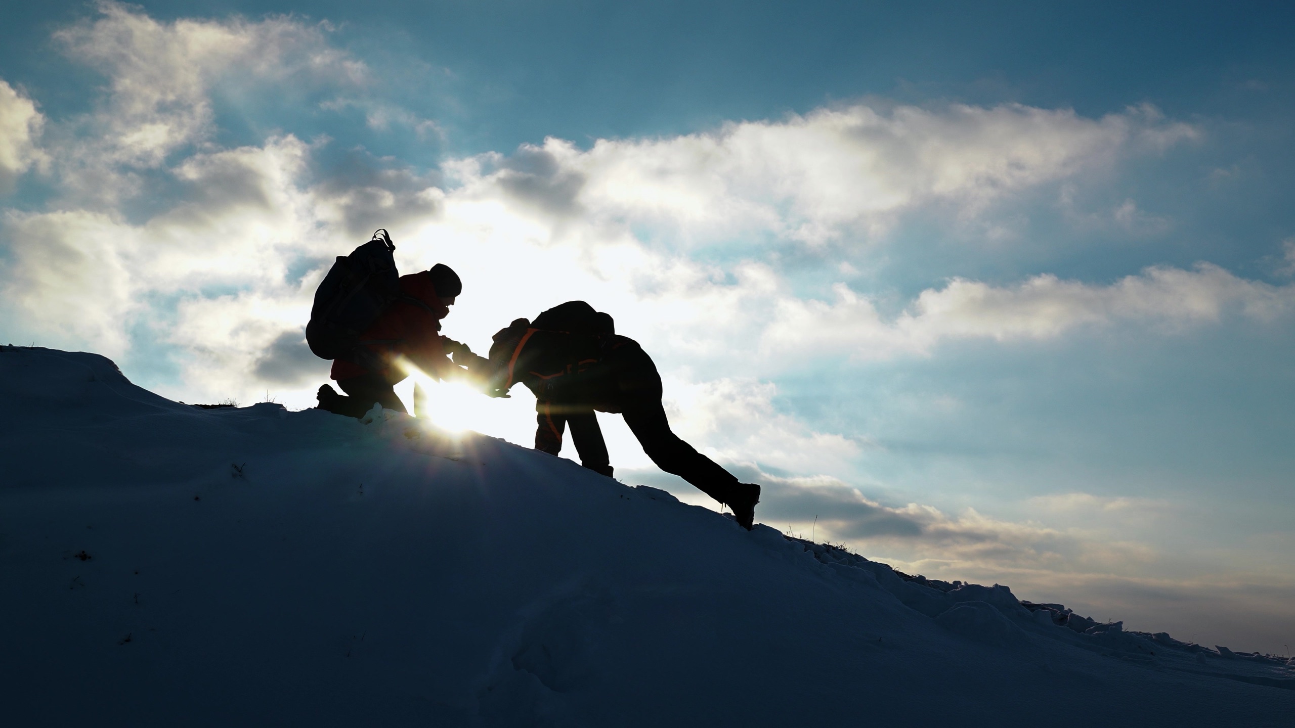 A climber reaching down to pull a teammate up a snowy ridge, silhouetted against the rising sun.