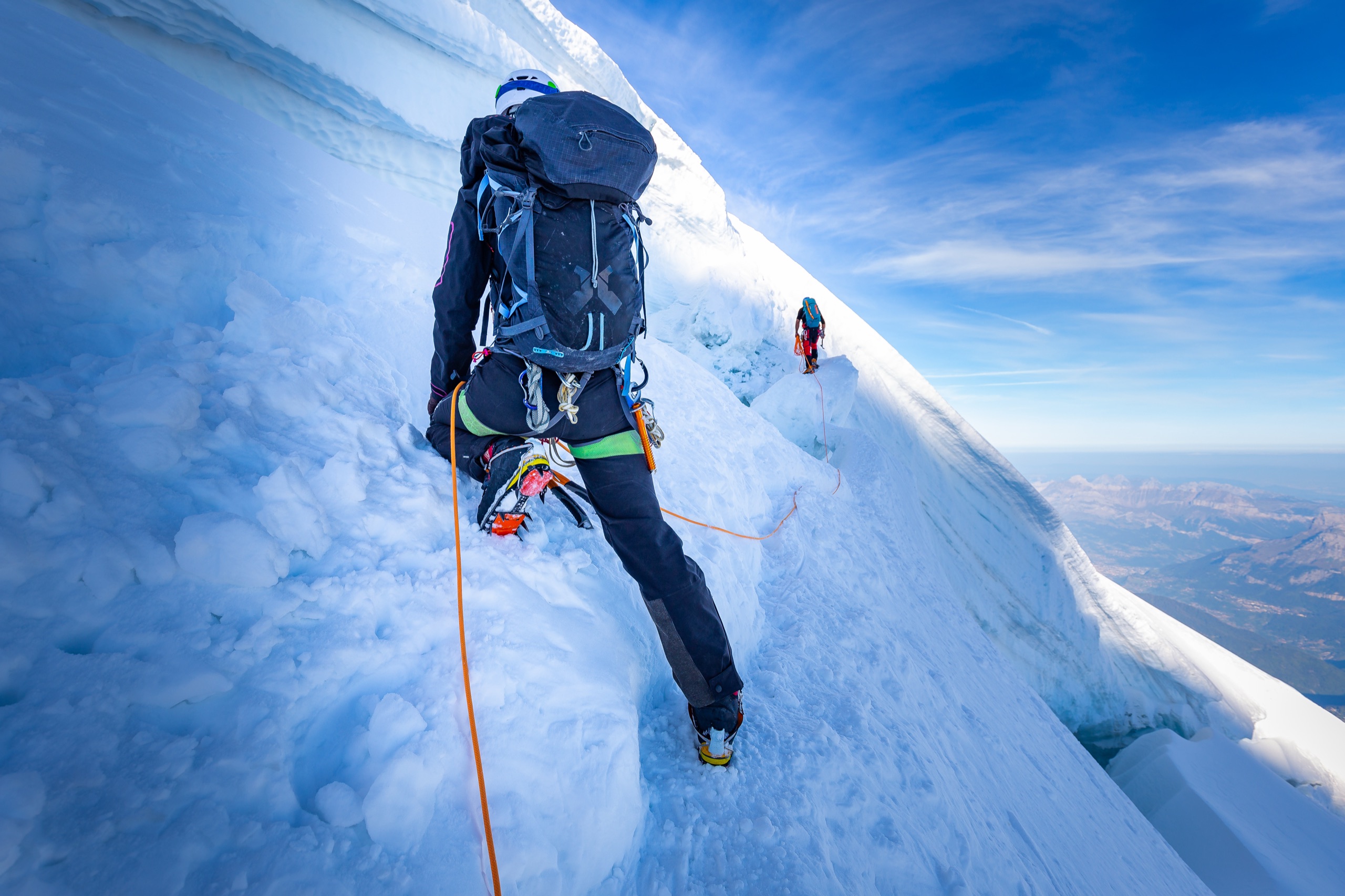 Two roped climbers with packs and ice axes ascending a steep glacier ridge high above the valleys.